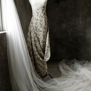 May include: A white and beige dress with a leaf pattern on the skirt. The dress is on a mannequin and is draped with a sheer white veil.