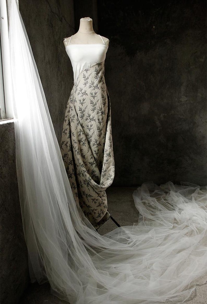 May include: A white and beige dress with a leaf pattern on the skirt. The dress is on a mannequin and is draped with a sheer white veil.