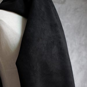 May include: A draped section of black fabric with a soft, velvety texture. The fabric is draped over a white form, with a gray background. The fabric appears to be a dark, solid color, possibly suede or velvet.