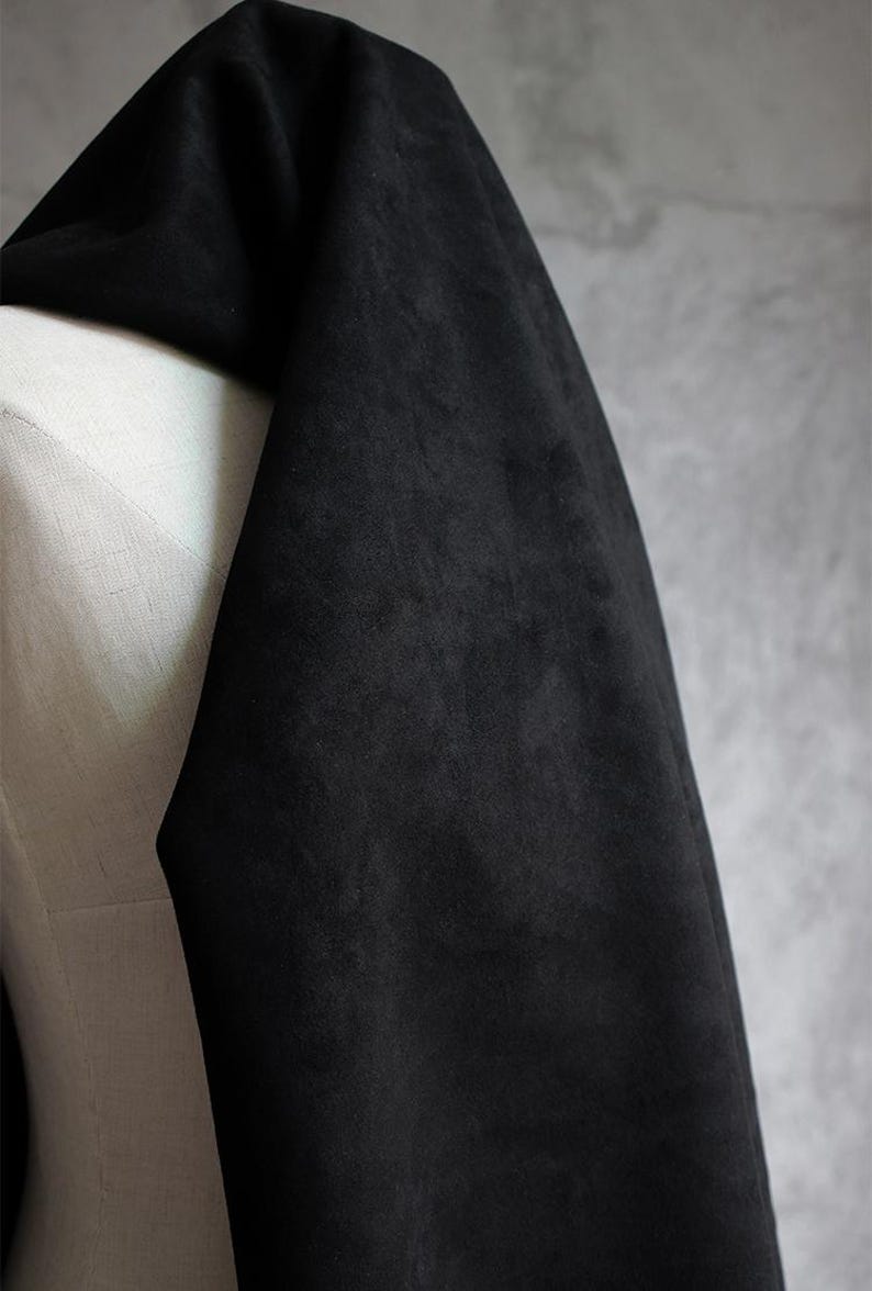 May include: A draped section of black fabric with a soft, velvety texture. The fabric is draped over a white form, with a gray background. The fabric appears to be a dark, solid color, possibly suede or velvet.