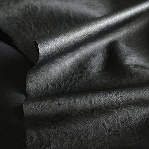 Op de afbeelding: Close-up van zwarte lederen stof met een getextureerd oppervlak. Het materiaal is gevouwen, waardoor de onderkant zichtbaar wordt en de glans van het leer wordt benadrukt. De afbeelding toont de soepele textuur en donkere kleur van het materiaal.