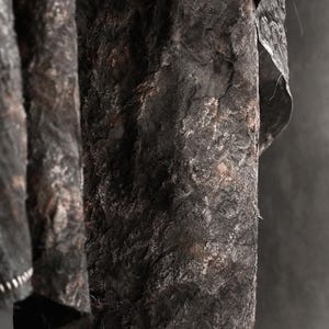 May include: Close-up of a textured, dark gray fabric with a rough, uneven surface. The fabric appears to be draped, with folds and shadows adding depth. The material has a mottled appearance with hints of brown.