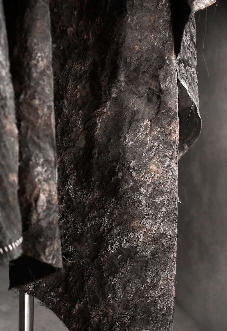 May include: Close-up of a textured, dark gray fabric with a rough, uneven surface. The fabric appears to be draped, with folds and shadows adding depth. The material has a mottled appearance with hints of brown.