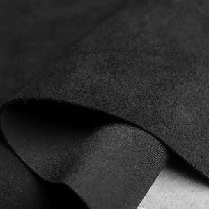 May include: Close-up of a folded piece of black fabric. The fabric has a soft, suede-like texture on the top layer and a smooth, tightly woven underside. The folds create shadows and depth, highlighting the material's texture and color.