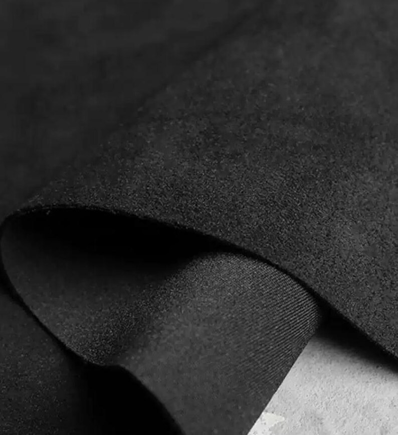 May include: Close-up of a folded piece of black fabric. The fabric has a soft, suede-like texture on the top layer and a smooth, tightly woven underside. The folds create shadows and depth, highlighting the material's texture and color.