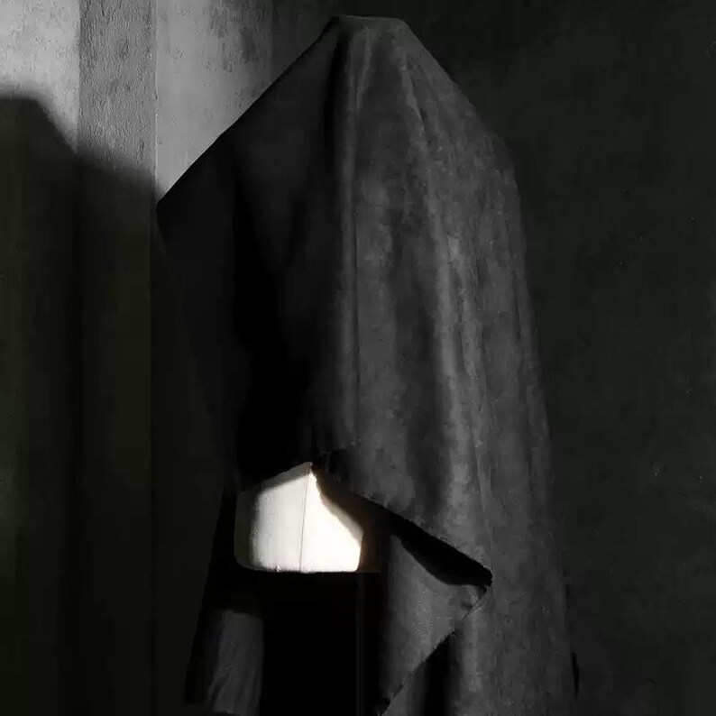 May include: A black fabric draped over a white dress form. The fabric has a rough texture and hangs in soft folds. The background is a dark gray, textured wall.