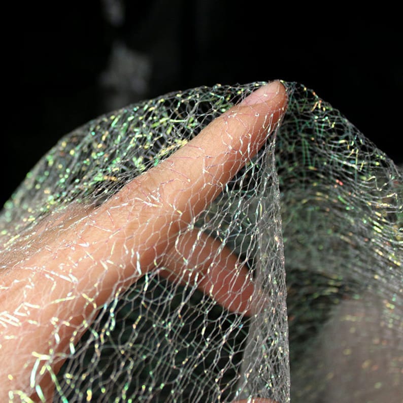 May include: Close-up of a shimmering, iridescent fabric with a net-like pattern. The fabric is held up, revealing its delicate texture and the way it catches the light, creating a sparkling effect. The background is dark, emphasising the fabric's luminosity.