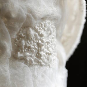 May include: Close-up of white fabric with a textured, raised design. The fabric appears to be layered, creating depth and dimension. The design is a focal point, adding visual interest to the soft, flowing material.