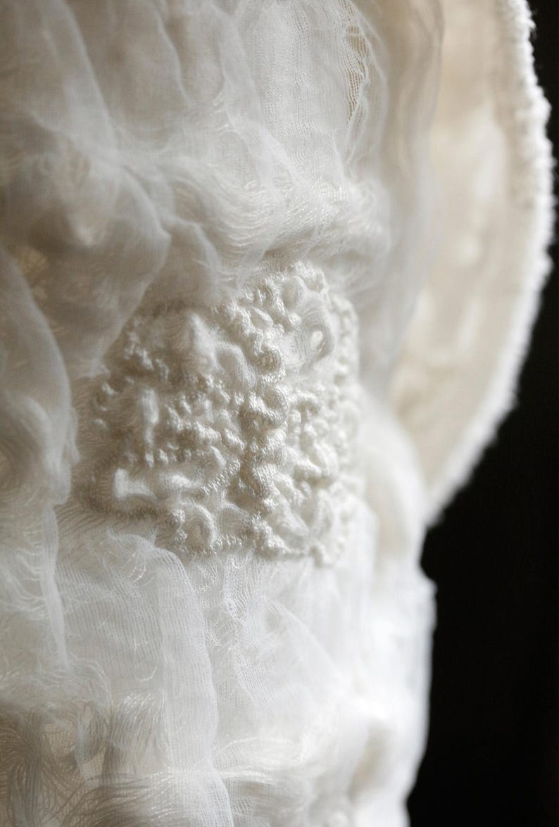 May include: Close-up of white fabric with a textured, raised design. The fabric appears to be layered, creating depth and dimension. The design is a focal point, adding visual interest to the soft, flowing material.