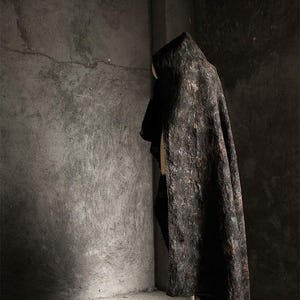 May include: A dark, textured cloak with a hood, displayed on a mannequin. The cloak has a mottled, bark-like appearance in shades of brown and black. The garment is draped, showcasing its shape and texture against a gray concrete background.