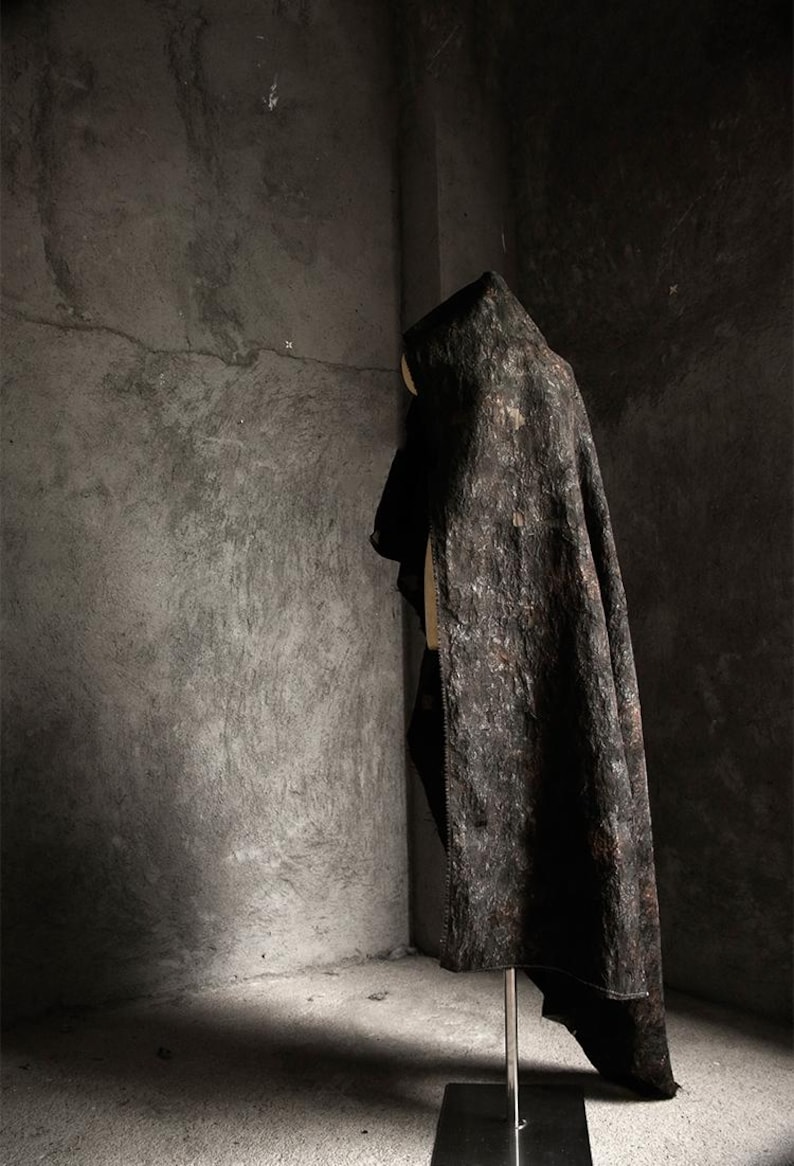 May include: A dark, textured cloak with a hood, displayed on a mannequin. The cloak has a mottled, bark-like appearance in shades of brown and black. The garment is draped, showcasing its shape and texture against a gray concrete background.