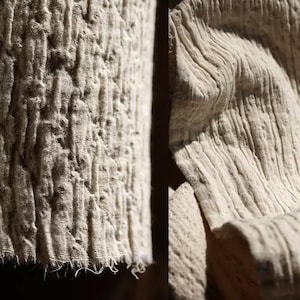 Op de afbeelding: Close-up van een getextureerde, gebroken witte stof. De linkerkant toont een gewatteerd patroon met verhoogde, onregelmatige bobbels. De rechterkant toont een geribbelde textuur met verticale lijnen en zachte plooien. De stof lijkt natuurlijk te zijn, mogelijk linnen of katoen.