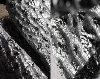 May include: Close-up of a silver fabric with a black pattern. The fabric has a textured, almost metallic, look.