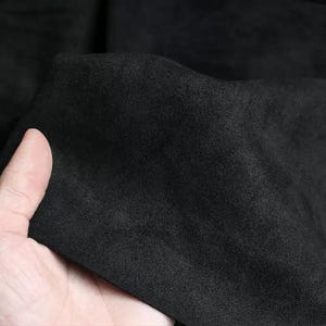 May include: Close-up of a piece of black fabric with a soft, suede-like texture. The fabric is held in a hand, showing its flexibility and the subtle variations in its surface. The material appears to be a solid, dark color.