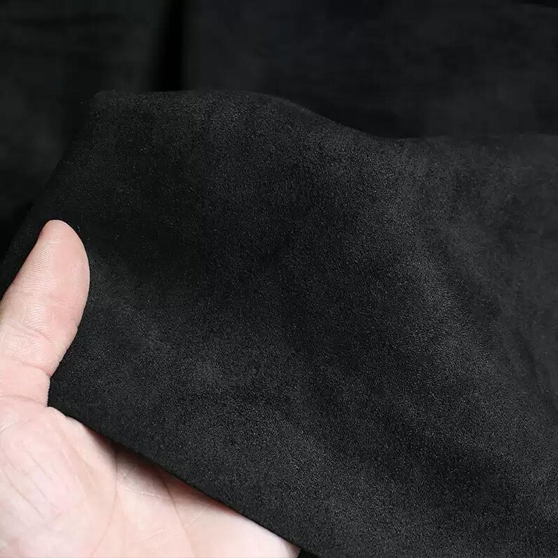May include: Close-up of a piece of black fabric with a soft, suede-like texture. The fabric is held in a hand, showing its flexibility and the subtle variations in its surface. The material appears to be a solid, dark color.
