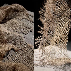 Puede incluir: Primer plano de una tela de yute tejida, de color marrón natural. La tela está dispuesta en pliegues suaves, con bordes deshilachados que añaden un aspecto rústico. El tejido grueso crea un patrón de rejilla, adecuado para proyectos de artesanía o decoración del hogar.