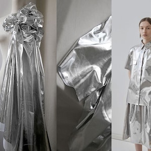Könnte beinhalten: Ein silberner Metallic-Stoff mit einer glänzenden, reflektierenden Oberfläche. Der Stoff ist drapiert und gefaltet und zeigt seine Textur und seinen Fall.