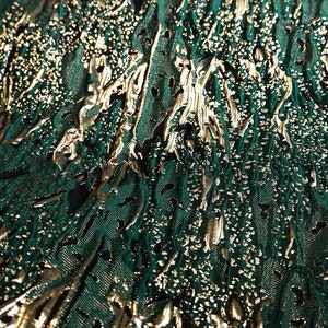 May include: Close-up of a green fabric with a gold metallic pattern. The fabric has a textured, embossed look.