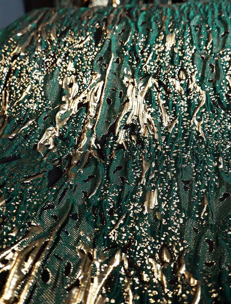 May include: Close-up of a green fabric with a gold metallic pattern. The fabric has a textured, embossed look.