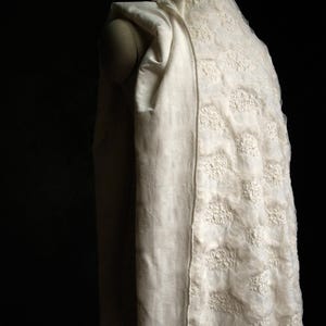 May include: A cream-colored, hooded garment with a textured floral pattern. The fabric drapes over a mannequin, showcasing the design and texture. The background is a deep black, highlighting the garment's details.