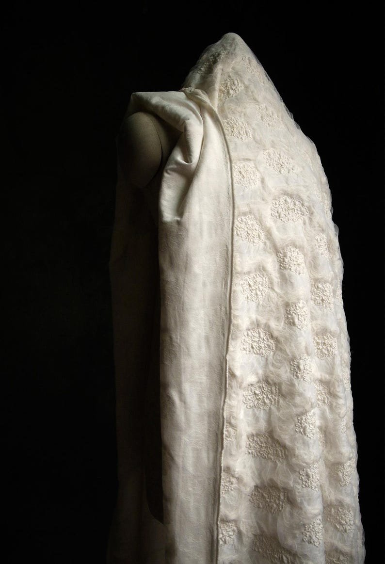 May include: A cream-colored, hooded garment with a textured floral pattern. The fabric drapes over a mannequin, showcasing the design and texture. The background is a deep black, highlighting the garment's details.