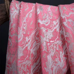 May include: Close-up of a pink fabric with a marbled design. The fabric has a swirling pattern of pink and white, creating a textured appearance. The fabric is draped, showing folds and shadows, and is likely used for clothing or home decor.