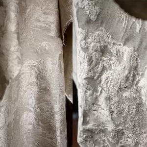 Könnte beinhalten: Nahaufnahme eines strukturierten, cremefarbenen Stoffes mit einem dezenten, erhabenen Blumenmuster. Der Stoff ist gefaltet und zeigt die Textur und die rohe Kante des Materials. Das Design hat einen leichten Glanz.