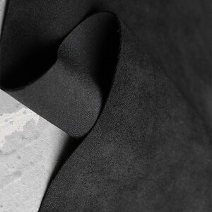 May include: Close-up of a black fabric with a soft, suede-like texture. The fabric is folded, revealing a contrasting smooth, woven underside. The image highlights the material's depth and tactile qualities, suitable for apparel or upholstery.