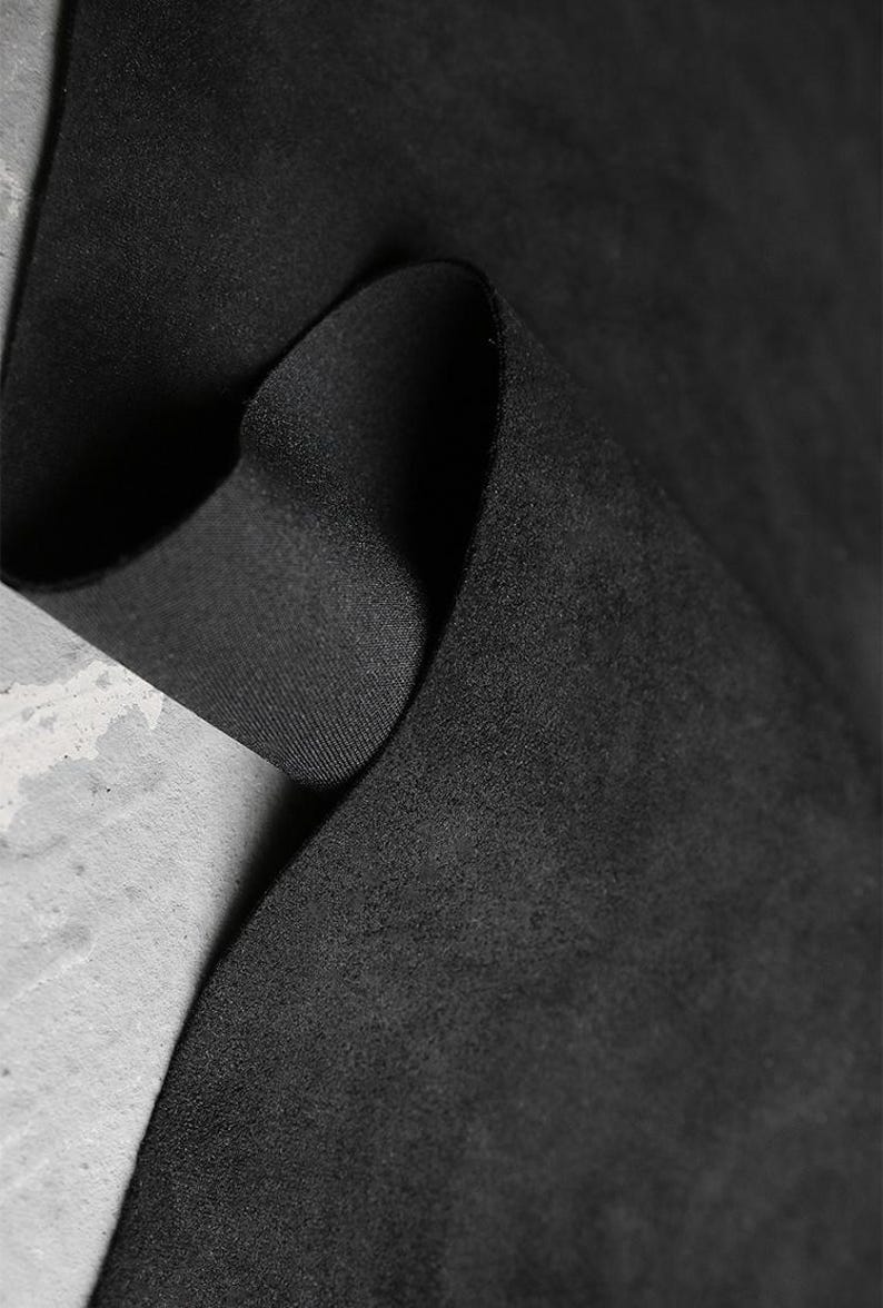 May include: Close-up of a black fabric with a soft, suede-like texture. The fabric is folded, revealing a contrasting smooth, woven underside. The image highlights the material's depth and tactile qualities, suitable for apparel or upholstery.