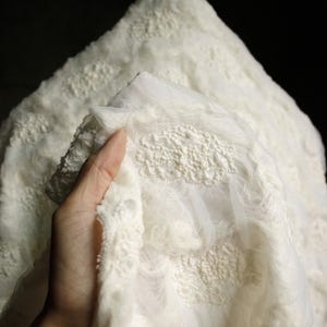 May include: Close-up of a white fabric with a textured, raised pattern. The fabric has a soft, fluffy appearance with a delicate, lacy overlay. The material is held up against a dark background, highlighting its intricate design.