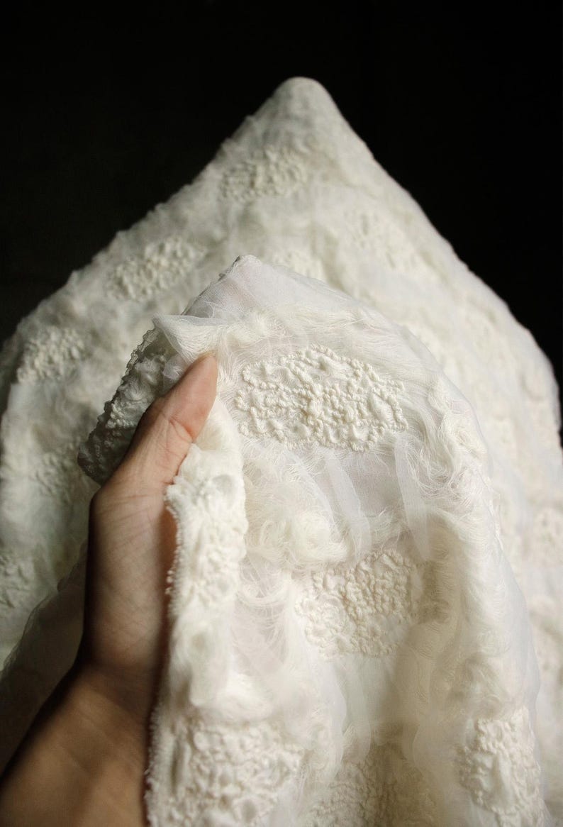 May include: Close-up of a white fabric with a textured, raised pattern. The fabric has a soft, fluffy appearance with a delicate, lacy overlay. The material is held up against a dark background, highlighting its intricate design.
