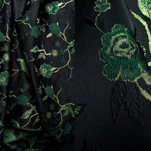 May include: Black fabric with an embroidered floral and bird design. The embroidery features green and gold thread, creating a detailed pattern of flowers, leaves, and birds against the dark background. The fabric appears to be a luxurious material.