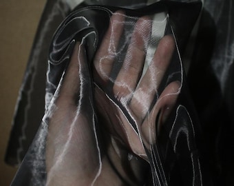 May include: Close-up of sheer black fabric with white highlights. The fabric is held up, revealing the texture and transparency. The material appears to be lightweight and flowing, suitable for apparel or decorative purposes.