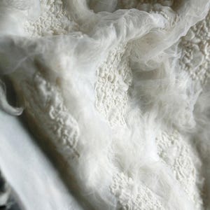 May include: Close-up of white fabric with a textured, layered design. The fabric appears to be a combination of delicate tulle and a more solid, possibly embroidered, material. The overall effect is soft and ethereal.