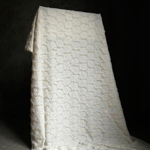 May include: A white, textured fabric draped over a rectangular form. The fabric has a repeating pattern of small, raised floral shapes. The background is dark, highlighting the bright white of the fabric.