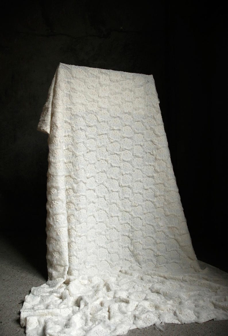 May include: A white, textured fabric draped over a rectangular form. The fabric has a repeating pattern of small, raised floral shapes. The background is dark, highlighting the bright white of the fabric.