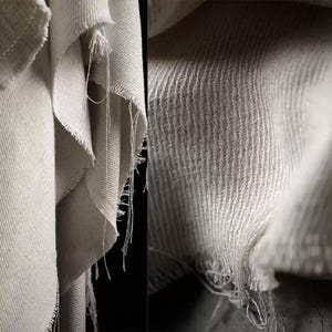 Puede incluir: Primer plano de tela blanquecina con tejido texturizado. La tela tiene bordes deshilachados e hilos sueltos, creando un aspecto crudo e inacabado. La imagen resalta la textura y el detalle del material.