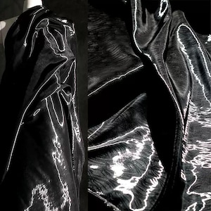 May include: Black garment with white accent lines, possibly a top or dress, draped on a mannequin. The fabric has a shiny, textured appearance. The design features a flowing, asymmetrical silhouette.