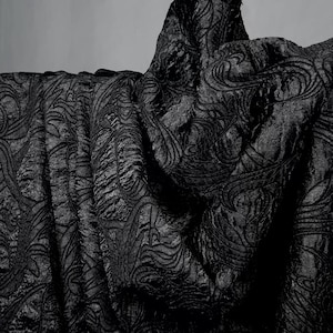 May include: Close-up of black fabric with an embossed, swirling pattern. The fabric is draped, creating folds and shadows that highlight the texture. The material appears thick and luxurious.