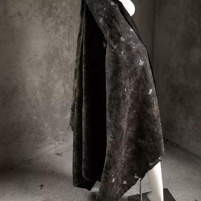 May include: A dark, textured fabric draped over a mannequin. The fabric appears to be a dark gray or black with lighter, irregular patches. The material hangs in soft folds, suggesting a garment or textile.