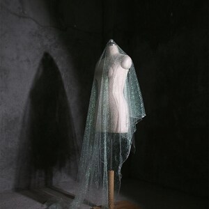 May include: A shimmering bridal veil with glitter is draped over a mannequin. The veil is a light, translucent colour, and it cascades down to the floor. The mannequin is on a wooden stand in a dimly lit room.