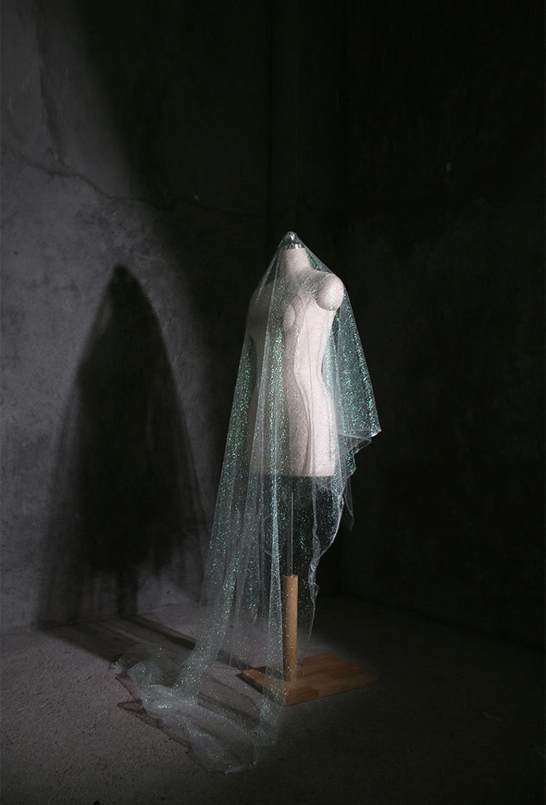 May include: A shimmering bridal veil with glitter is draped over a mannequin. The veil is a light, translucent colour, and it cascades down to the floor. The mannequin is on a wooden stand in a dimly lit room.