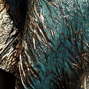 May include: Close-up of a fabric with a textured, metallic gold and teal green pattern. The fabric has a woven, almost shimmering appearance.
