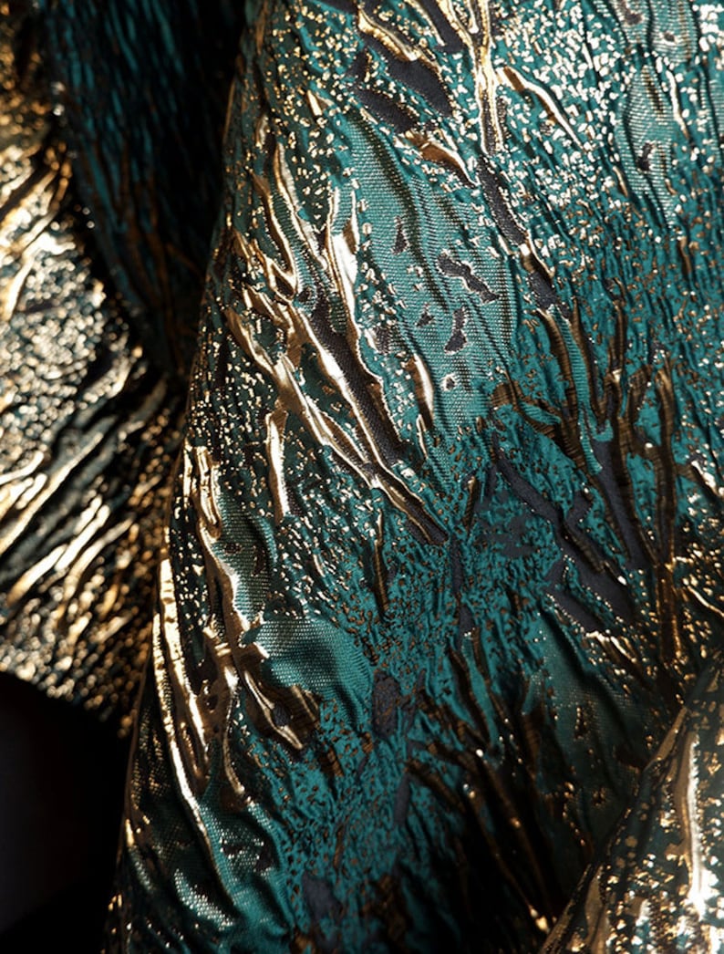 May include: Close-up of a fabric with a textured, metallic gold and teal green pattern. The fabric has a woven, almost shimmering appearance.