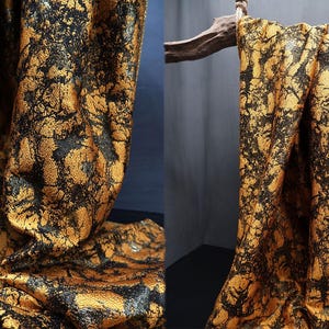 Op de afbeelding: Close-up van een stof met een goud en zwart patroon. De stof is gedrapeerd en toont plooien en textuur. Het ontwerp heeft een gevlekt uiterlijk, met zwarte vormen tegen een gouden achtergrond. De stof wordt waarschijnlijk gebruikt voor kleding of woondecoratie.