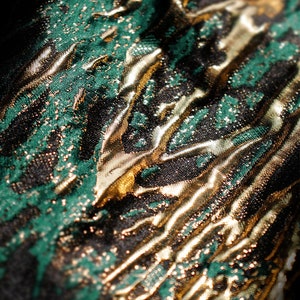 May include: Close-up of a green and gold fabric with a textured, metallic finish. The fabric has a swirling pattern and a shimmering effect.