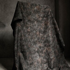 May include: A dark, textured fabric draped over an unseen form. The fabric has a mottled appearance with shades of black, brown, and gray. The folds and draping create shadows, adding depth and dimension to the textile.