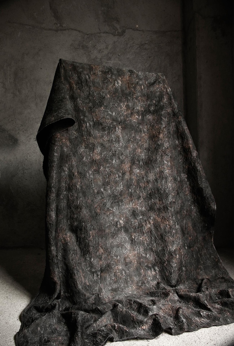 May include: A dark, textured fabric draped over an unseen form. The fabric has a mottled appearance with shades of black, brown, and gray. The folds and draping create shadows, adding depth and dimension to the textile.