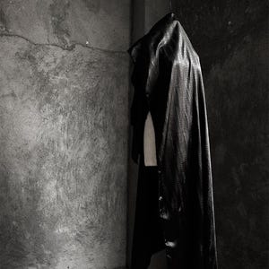 May include: A black satin cape draped over a mannequin in a dimly lit room with a concrete wall in the background.