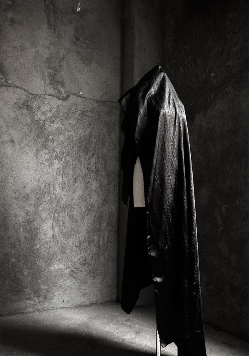 May include: A black satin cape draped over a mannequin in a dimly lit room with a concrete wall in the background.